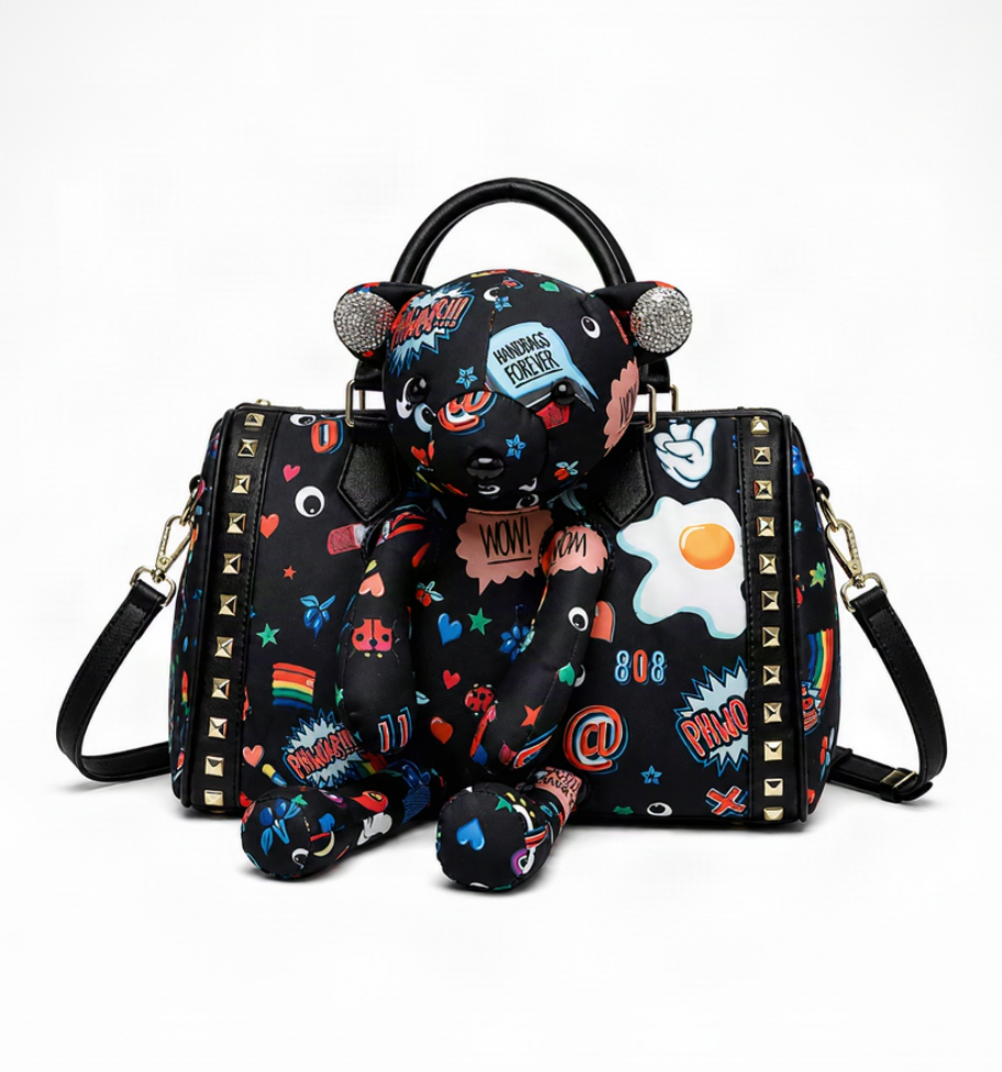 A black duffle-style handbag with a plush bear character attached on top, featuring a colorful all-over graphic print with cartoon eggs, hearts, stars, and comic-style text. The bag has black handles, a detachable shoulder strap, and gold-tone studs along the side.