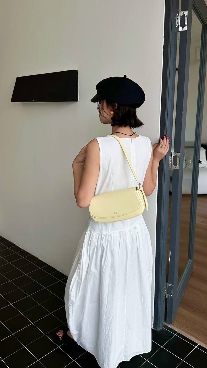 A woman wearing a white sleeveless midi dress, a black beret, and black sandals is shown from behind, carrying a pale yellow crescent-shaped shoulder bag with a thin strap.