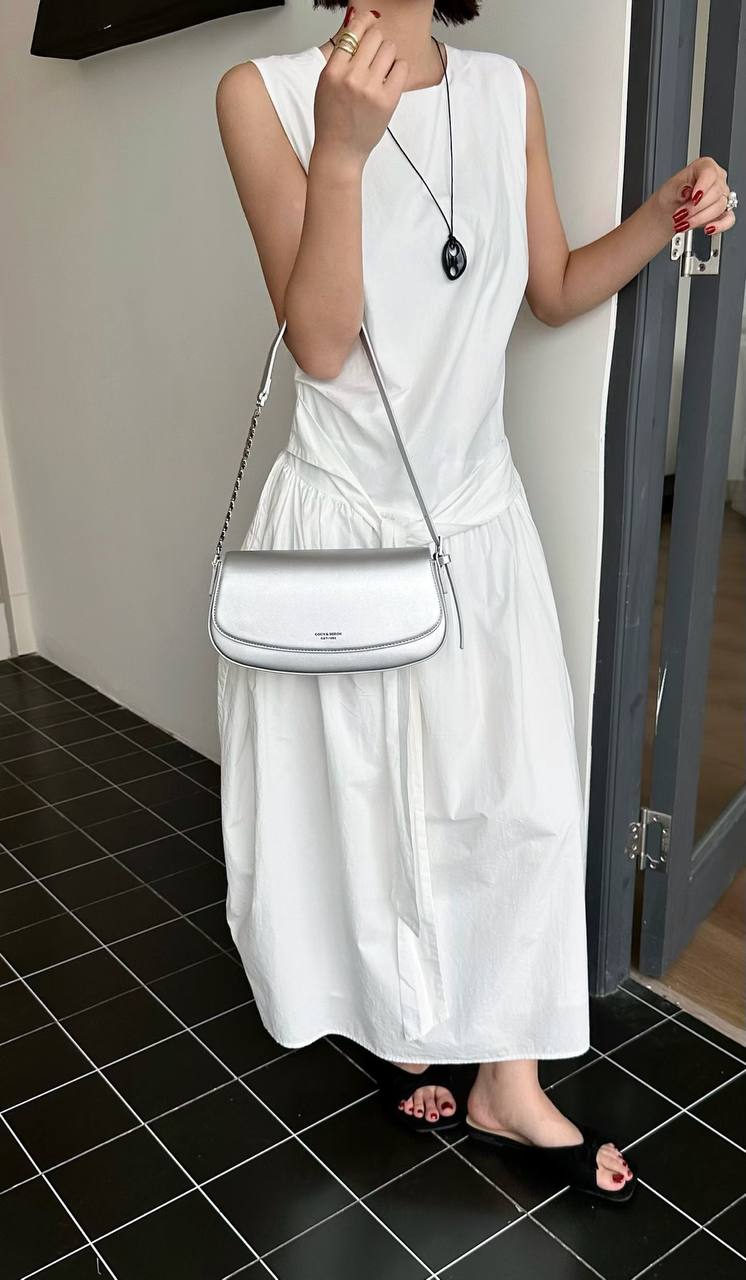 A woman wearing a white sleeveless maxi dress and black slide sandals, carrying a white leather shoulder bag with a silver chain strap in an indoor setting with black tiled flooring.