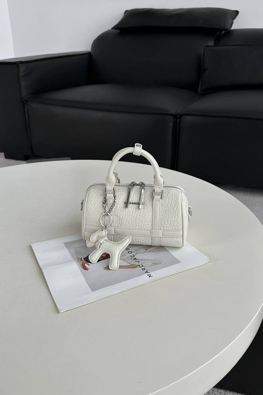 A small structured handbag in textured off-white leather sits on a round white table, with a black sofa in the background. The bag has top handles, a chain detail, and a small hanging charm.
