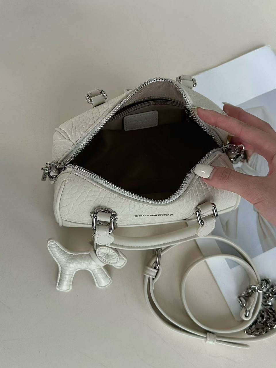 A white textured leather handbag with silver hardware, shown open with a hand resting near the zipper. A matching white dog-shaped charm hangs from the handle, and a coiled strap with a silver chain detail lies beside it on a light beige surface.