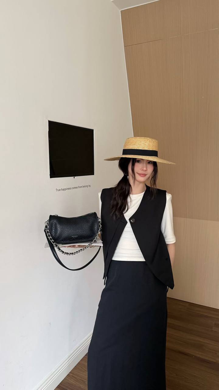 A woman wearing a straw hat with a black band, a black sleeveless vest over a white top, and a long black skirt, standing indoors next to a black shoulder bag hanging on a wall.