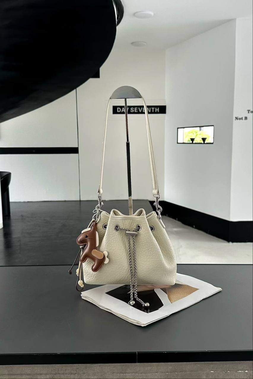 A cream-colored textured leather bucket bag with a brown leather monkey charm, displayed on a stand on top of a magazine in a modern room setting.