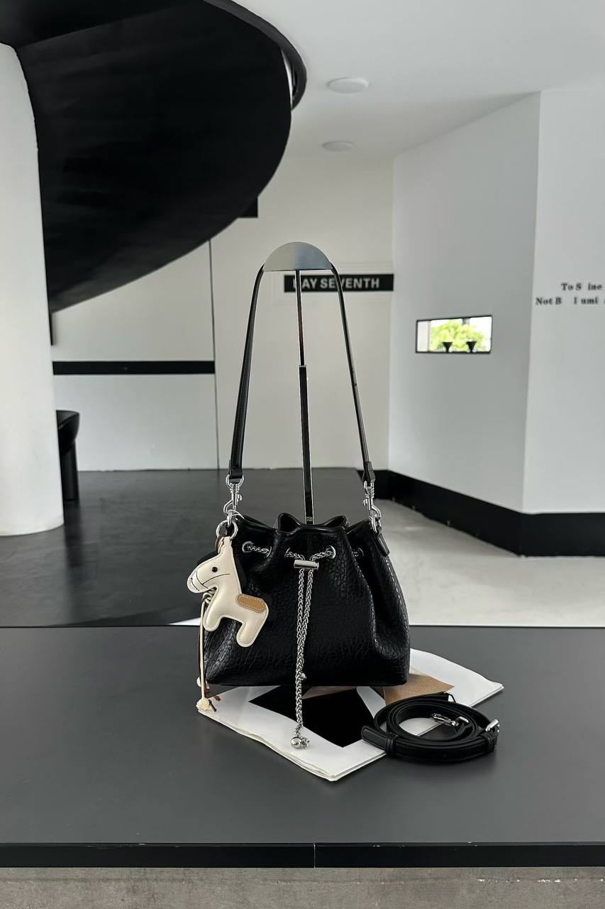 A black leather bucket bag with a drawstring closure, displayed on a black table in a modern interior setting. The bag features a black shoulder strap, a silver chain detail, a small white leather horse charm, and a matching black ring strap.