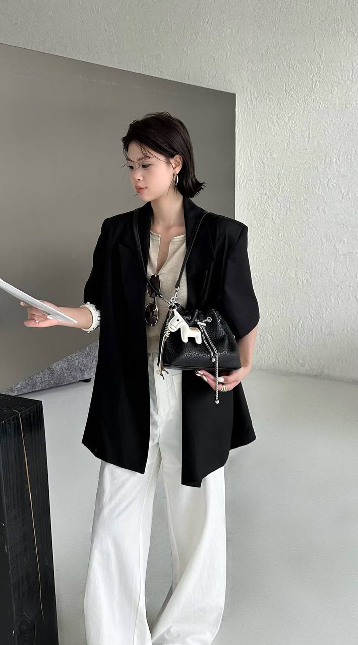 A woman stands indoors wearing a black blazer over a beige top and white wide-leg pants, holding a black handbag with a white horse charm. She is posed in a minimalist room with textured walls.