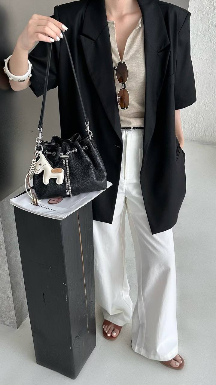 A person wearing a black short-sleeve blazer, white wide-leg pants, and a beige top carries a black textured bucket bag with a silver chain strap and a white horse charm. The person is standing next to a black display block in a neutral indoor setting.