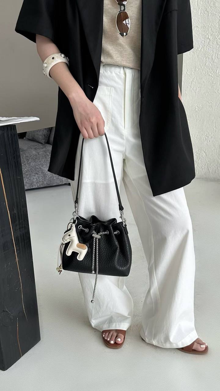 A person wearing a black blazer, beige top, and white wide-leg pants holds a small black bucket bag with a long strap and dangling charm in an indoor setting.