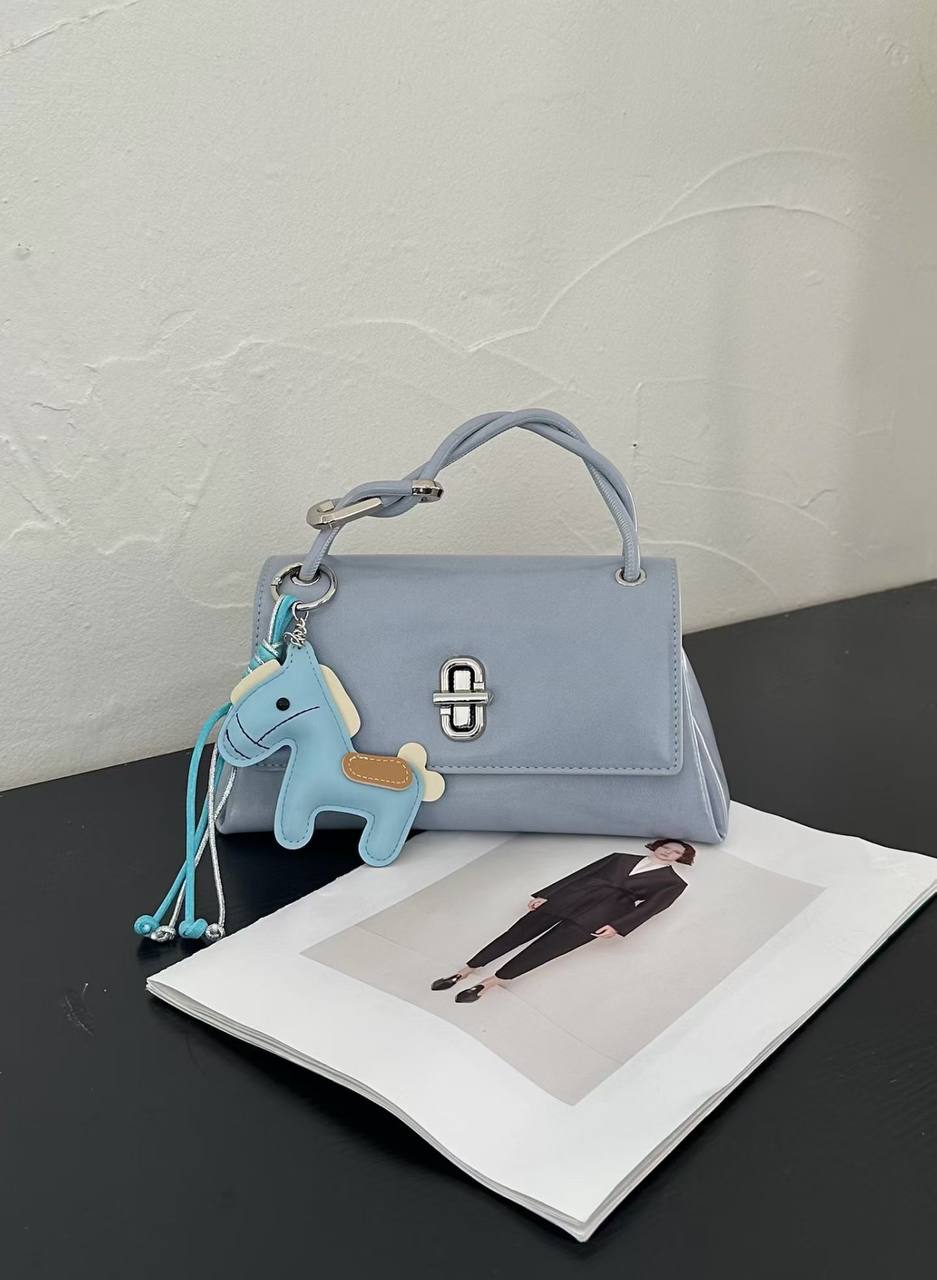 A light blue structured handbag with a top handle and silver turn-lock clasp, styled on a dark tabletop with an open magazine. A small blue horse-shaped keychain charm with a tassel is attached to the bag.