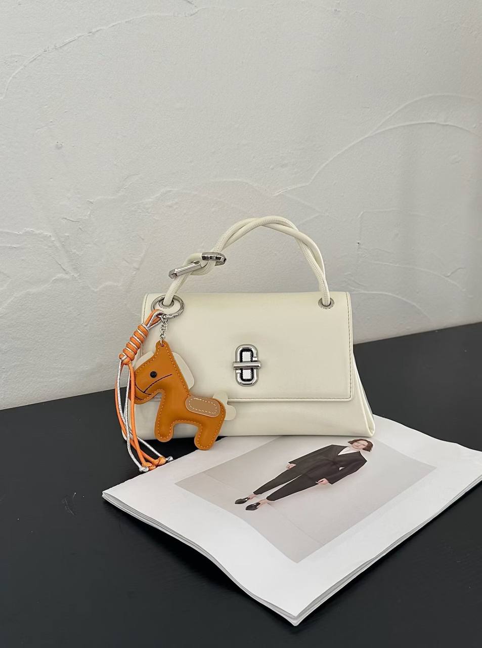 White structured top handle handbag with silver twist-lock closure and decorative orange horse charm on a white studio background.