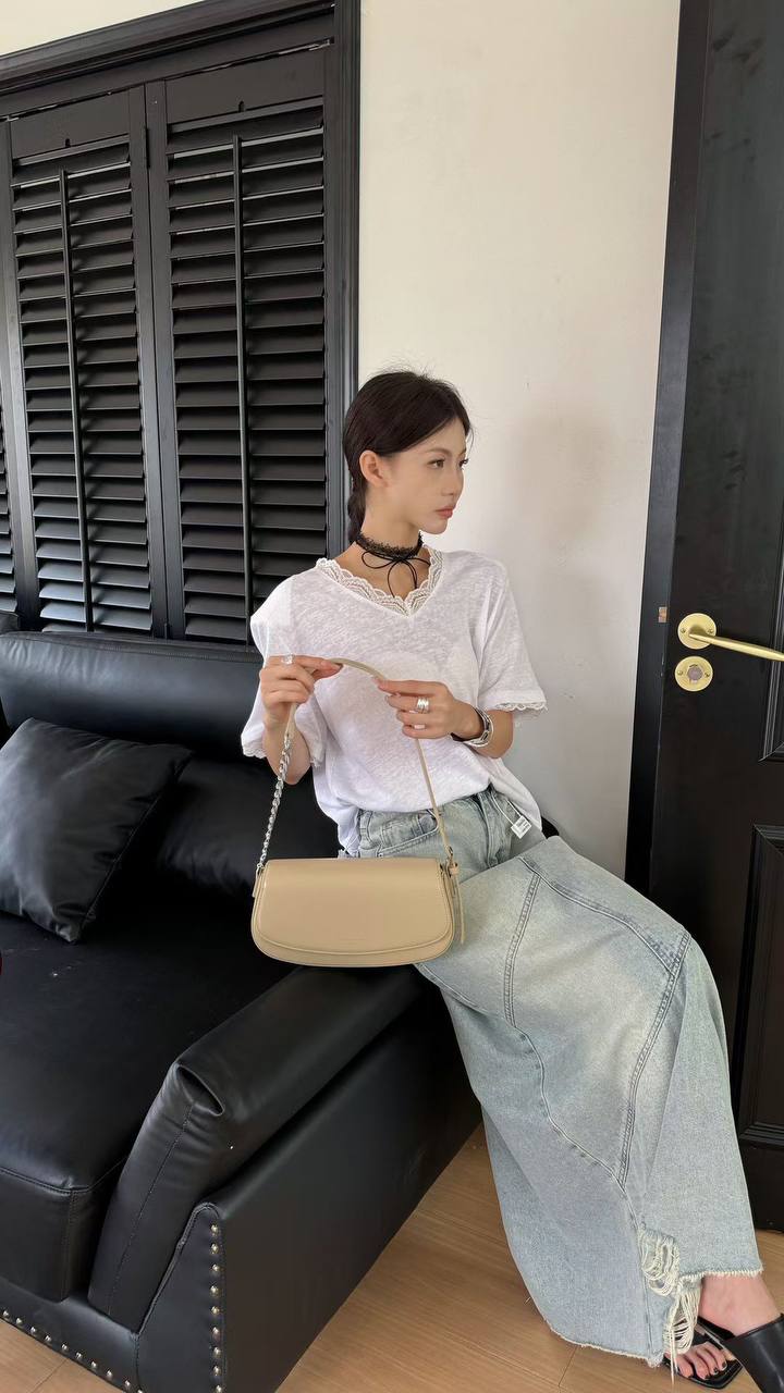 A woman sits on a black leather sofa wearing a white short-sleeve blouse, light-wash wide-leg denim jeans, and black sandals, while carrying a beige shoulder bag.