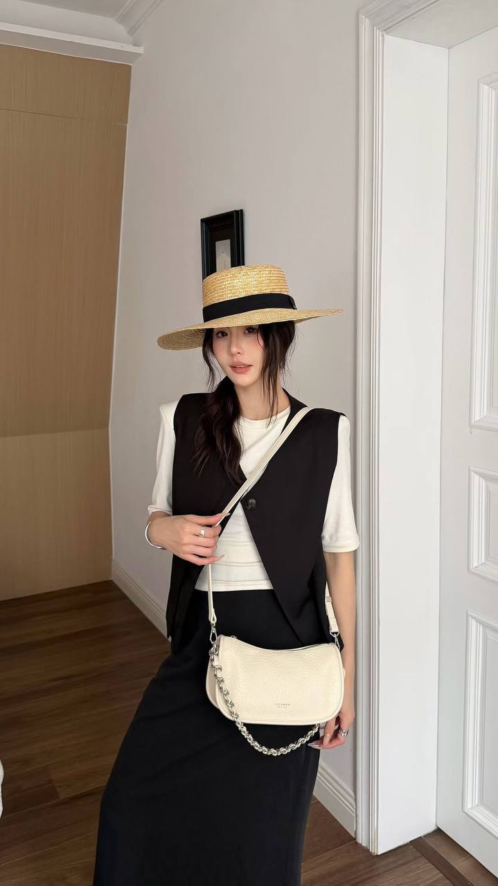 A woman wearing a straw hat with a black band, a black vest over a white short-sleeve top, a black skirt, and carrying a cream-colored shoulder bag with a chain strap, standing in an indoor room with white walls and wooden floors.