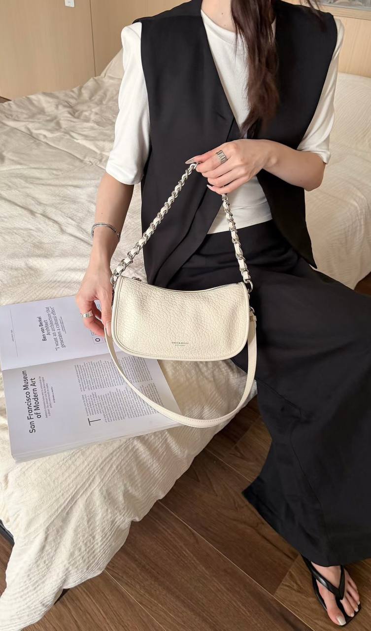 A woman wearing a black vest and white shirt sits on a bed holding a cream-colored pebbled leather shoulder bag with a silver chain strap.