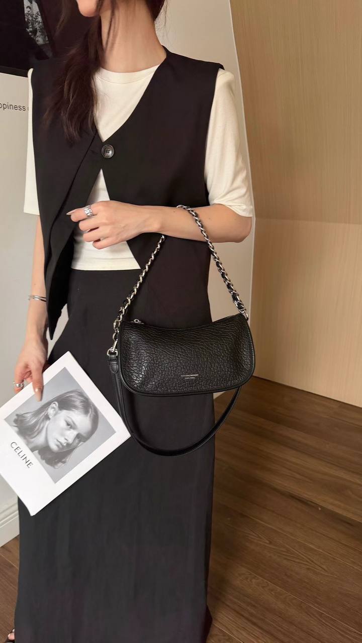 A woman models a black crocodile-embossed shoulder bag with a silver chain strap, worn crossbody over a black vest and white t-shirt.