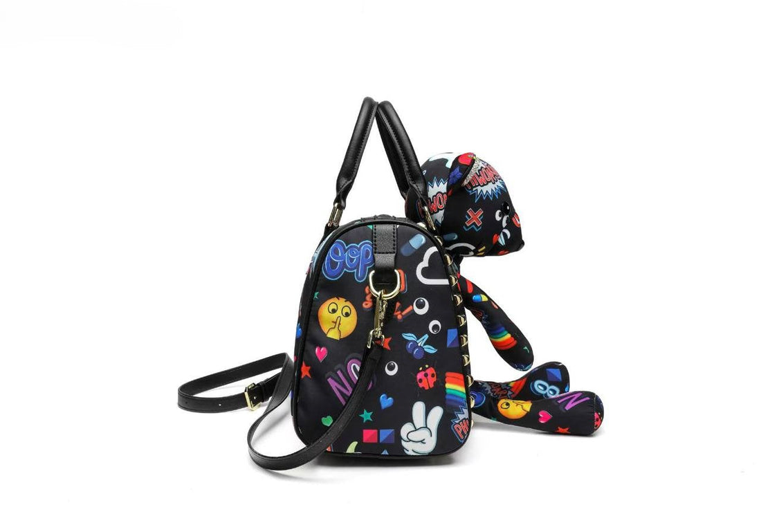 A black duffle-style handbag with a plush bear character attached on top, featuring a colorful all-over graphic print with cartoon eggs, hearts, stars, and comic-style text. The bag has black handles, a detachable shoulder strap, and gold-tone studs along the side.